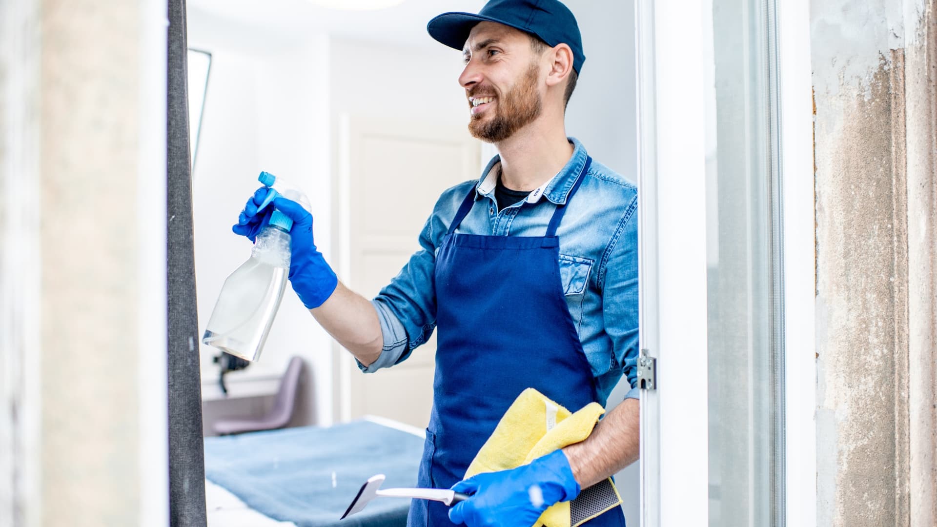 Male cleaner in an apron holding a spray bottle, microfiber cloth, and smiling. 