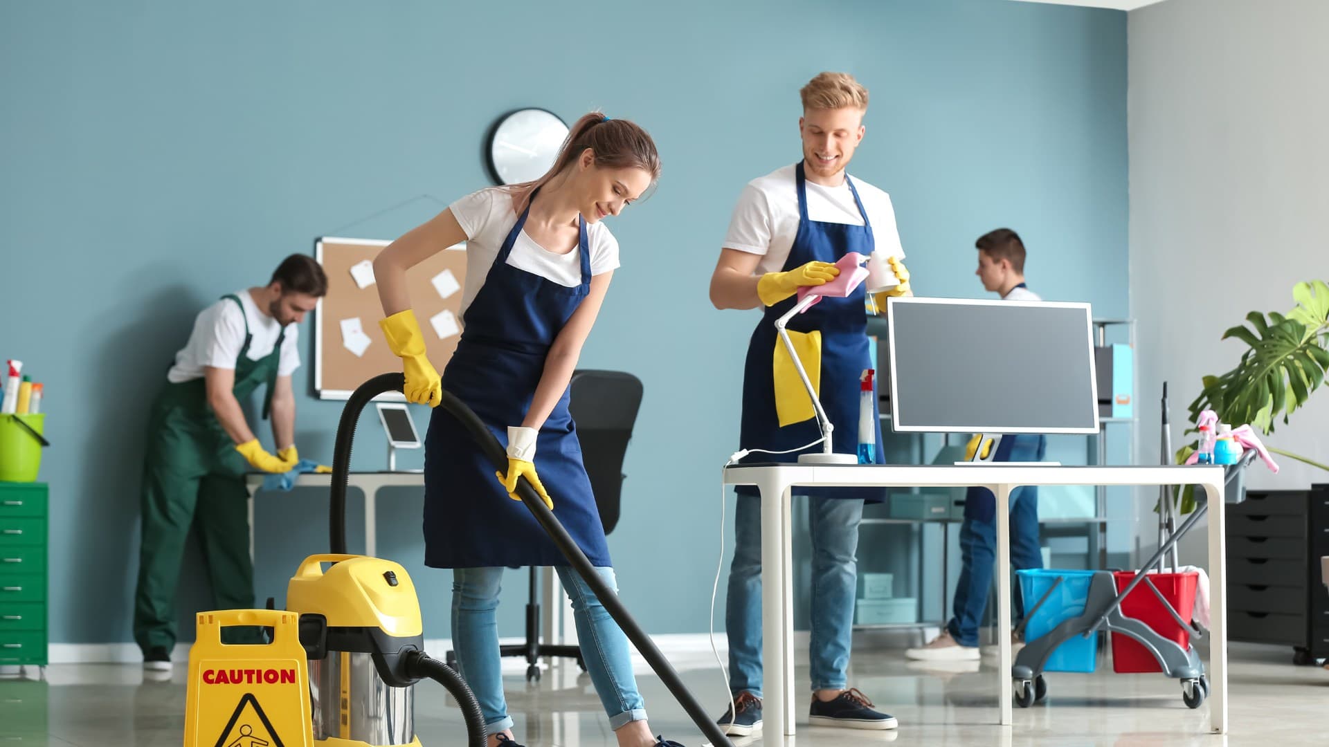 Cleaning crew of four in a commercial office room vacuuming, dusting, and sanitizing 