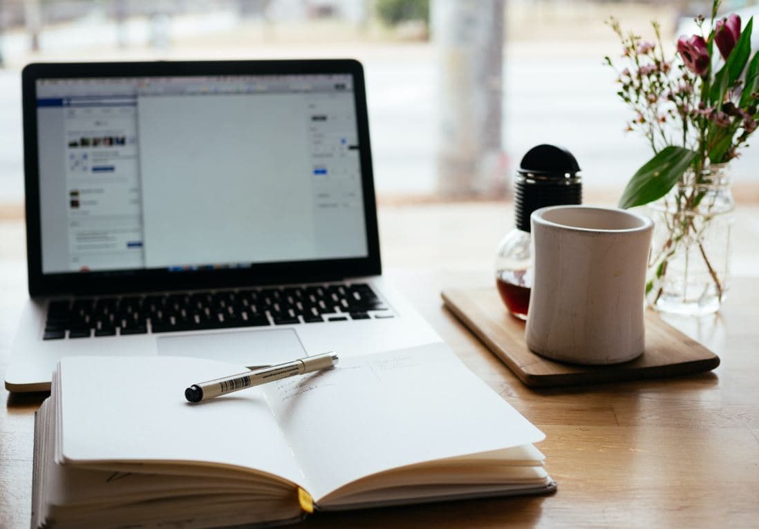 Open laptop with a notebook and pen before the laptop and a coffee mug to the right 